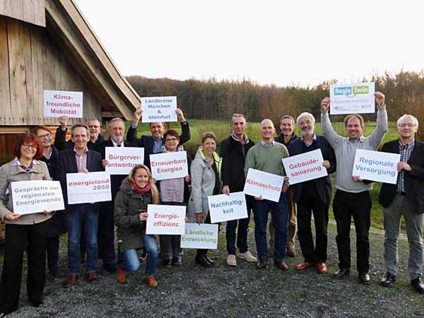 Foto: Vertreter der Landkreise München und Steinfurt stehen vor einem Ziegelsteingebäude und halten Schilder in die Luft, auf denen Stichworte rund um die Themen "Energie" und "Klimaschutz" zu lesen sind.