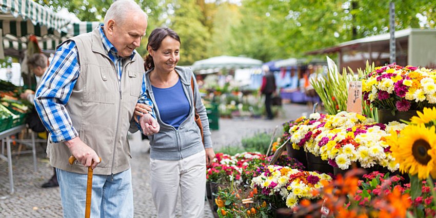 Eine Frau führt einen älteren, am Stock gehenden Mann über einen Markt. Foto: istockphoto.com/FredFroese