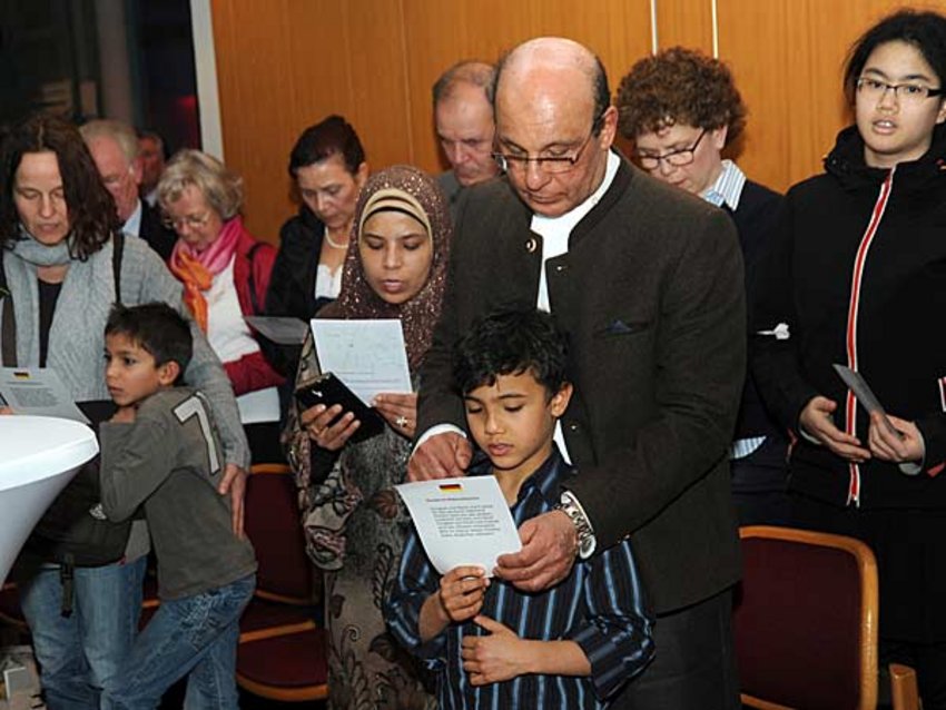 Foto: Die Gäste des Empfangs für Neueingebürgerte halten die bereitgestellten Liedzettel mit den Texten der deutschen Nationalhymne und der Bayernhymne in den Händen und singen.